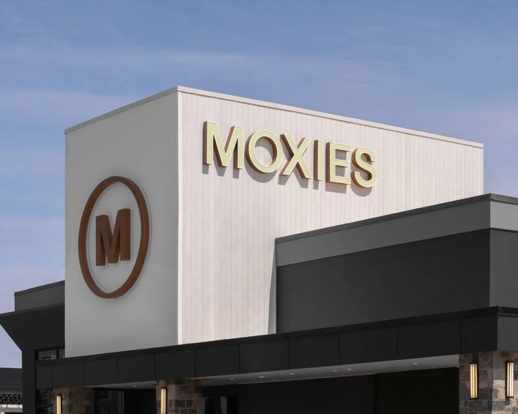 Modern commercial building exterior featuring vertical white custom aluminum siding, a large gold MOXIES sign, and a brown logo.