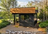 Modern black shed with textured aluminum siding used as a backyard bar, situated on a grassy lawn near tall trees.